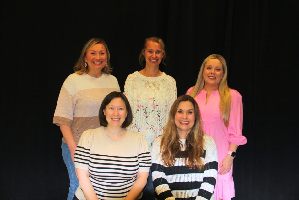 Group portrait of five women smiling in front of a black backdrop.